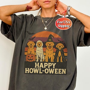 Puede incluir: Camiseta gris oscuro con un diseño temático de Halloween. El diseño presenta cuatro perros golden retriever disfrazados de calabaza, momia, bruja y vampiro, con el texto "HAPPY HOWL-OWEEN" debajo. Una silueta de murciélago está encima de los perros.