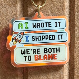May include: A rectangular acrylic keychain with a gold-tone metal clasp topped by a white and yellow daisy charm. The keychain features pixelated text reading AI WROTE IT, I SHIPPED IT, and WE'RE BOTH TO BLAME, accompanied by a small rocket ship graphic.