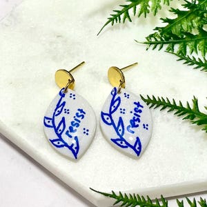 May include: A pair of white and blue earrings with a gold post. The earrings are teardrop-shaped with a blue floral design and the word "resist" written in blue. The earrings are on a white marble surface.