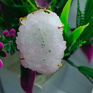 Puede incluir: Un colgante de oro con una piedra rosa claro texturizada con manchas verdes y blancas. El colgante tiene una fianza dorada y una piedra púrpura más pequeña. El fondo incluye follaje verde y púrpura.