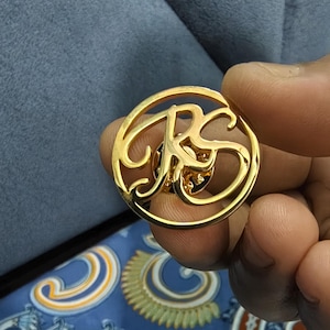 Peut inclure: Épinglette dorée avec les lettres entrelacées "RS" dans un cercle. L'épinglette est tenue sur un fond bleu, suggérant qu'elle pourrait être portée sur des vêtements ou des accessoires. La surface de l'épinglette est brillante et réfléchissante.