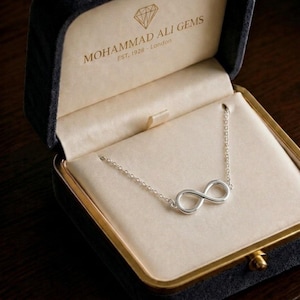Op de afbeelding: Een zilveren ketting met een oneindigheidssymbool, gepresenteerd in een open zwarte sieradendoos met een crèmekleurig interieur. De ketting heeft een fijne ketting en een oneindigheidshanger. De doos heeft gouden randen en de tekst "MOHAMMAD ALI GEMS EST. 1928 - London".