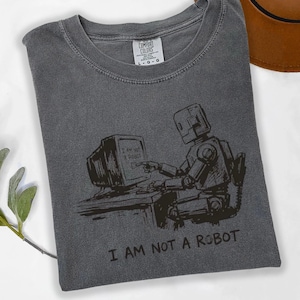 May include: A charcoal gray t-shirt featuring a black graphic of a robot sitting at a desk, interacting with a computer monitor. The screen displays the text I AM NOT A ROBOT, which is also printed in large capital letters below the illustration.