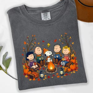 May include: A charcoal gray t-shirt featuring a graphic print of the Peanuts characters sitting around a campfire in an autumn forest. The design includes Snoopy, Charlie Brown, Lucy, Linus, and Sally surrounded by orange leaves and pumpkins.