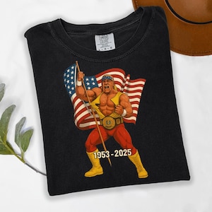 May include: Black t-shirt featuring a cartoon illustration of a wrestler holding an American flag. The wrestler is wearing red pants, yellow boots, and a championship belt. The text "1953-2025" is below the wrestler.