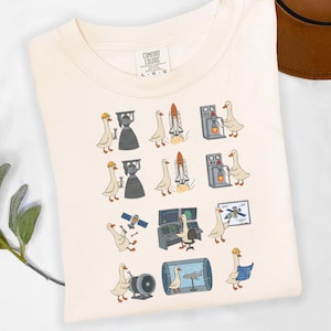 May include: A cream-colored t-shirt featuring a graphic print of white geese performing various aerospace engineering tasks, including working on rocket engines, launching space shuttles, operating control consoles, and reviewing technical blueprints.