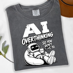 May include: A charcoal gray crew neck t-shirt featuring a white graphic of a robot sitting in a beanbag chair holding a mug. The text reads AI OVERTHINKING SO YOU DON'T HAVE TO. The shirt has a Comfort Colors brand label at the inner neckline.