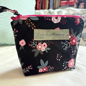 Op de afbeelding: Zwarte etui met bloemenprint, roze rits en een stoffen label met "HOUSE OF HOLBROOK". De etui heeft roze en witte rozen, groene bladeren en kleine oranje accenten.