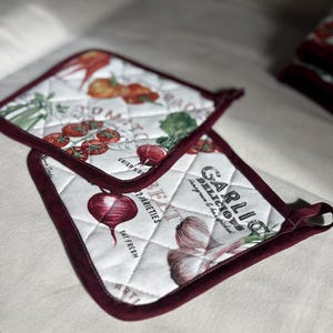 Könnte beinhalten: Zwei gesteppte Topflappen mit weißem Hintergrund und bordeauxfarbenem Rand. Einer zeigt Tomaten und Karotten, der andere Knoblauch und Rüben. Der Text enthält "GARLIC DELICIOUS" und "EAT FRESH".