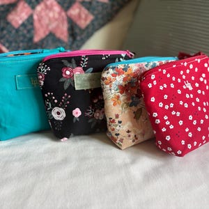 Pode incluir: Quatro pequenas bolsas com zíper em várias cores e padrões. Uma é turquesa, outra preta com estampa floral, uma terceira com um padrão floral em um fundo bege e a última vermelha com detalhes florais brancos.