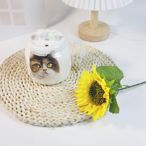 以下が含まれることがあります： 猫の顔のデザインが施された白いセラミック製のペット用ウォーターファウンテン。 織られたマットの上に噴水があり、黄色のヒマワリの隣にあります。 白いシェードの小さなランプが背景にあります。