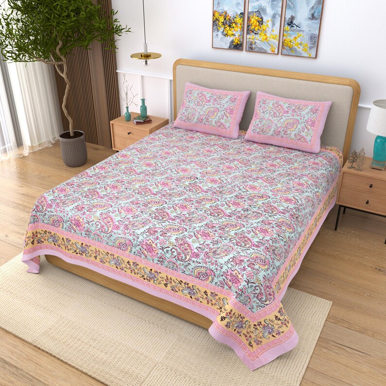 May include: A floral patterned bedspread and two pillowcases in pink, yellow, and blue hues. The bedspread features a repeating paisley design with a yellow border. The pillows have a pink border. The bed is made up with a light-colored rug underneath.