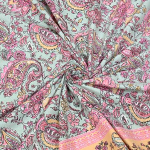 May include: A close-up of a fabric with a paisley pattern. The fabric has a light green background with pink, peach, and yellow floral and paisley designs. The fabric is folded and draped, showing the intricate details of the pattern.