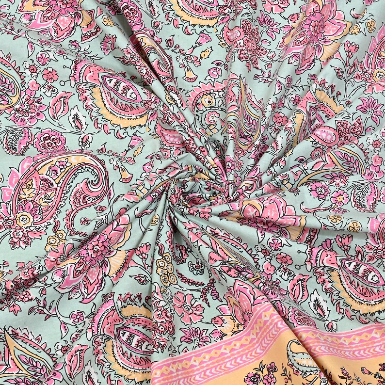 May include: A close-up of a fabric with a paisley pattern. The fabric has a light green background with pink, peach, and yellow floral and paisley designs. The fabric is folded and draped, showing the intricate details of the pattern.