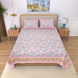 Op de afbeelding: Een beddenset met een paisley patroon in roze, geel en paars op een lichtblauwe achtergrond. De set bevat een laken en twee kussenslopen met roze randen. Het bed is opgemaakt met de lakens en kussenslopen.