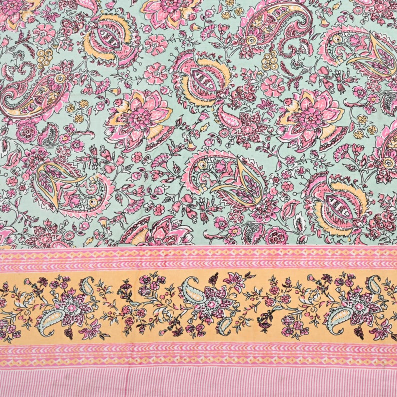 May include: A floral textile featuring a paisley and floral pattern in shades of pink, yellow, and green. The design includes a border with paisley and floral motifs on a yellow background, and a striped pink bottom.