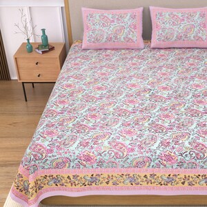 May include: A bed set featuring a light blue paisley pattern with pink and yellow accents. The set includes a patterned bedspread and two matching pillowcases with pink borders. The bedding is displayed on a wooden bed frame.