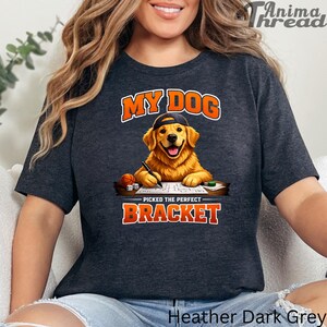 May include: Heather dark grey t-shirt with the text "MY DOG PICKED THE PERFECT BRACKET". The graphic features a golden retriever wearing a cap and writing on a paper, with a basketball and dice. The shirt is a comfortable, casual wear.