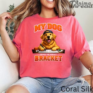 May include: Coral-colored t-shirt with a graphic of a Golden Retriever wearing a cap and writing on a notepad. The text above the dog reads "MY DOG" and below it, "PICKED THE PERFECT BRACKET".