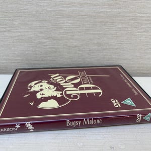 Può includere: Una custodia DVD bordeaux per il film "Bugsy Malone". La custodia presenta una grafica di due persone e il titolo del film. La custodia del DVD è di dimensioni standard e presenta il logo Carlton.