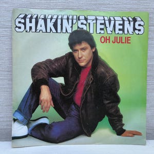 May include: Album cover for Shakin' Stevens' "Oh Julie." The cover shows a man in a brown leather jacket, red shirt, and blue jeans. The album title is in a white, stylized font against a green background.