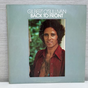 Puede incluir: Portada del álbum "Back to Front" de Gilbert O'Sullivan. La portada presenta un retrato de un hombre con cabello oscuro y rizado y una camisa roja. El título del álbum y el nombre del artista están en blanco en la parte superior.