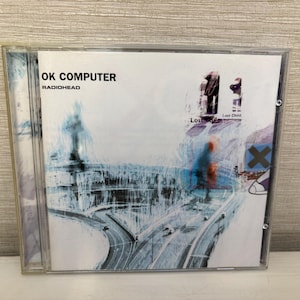 May include: A Radiohead OK Computer CD in a clear plastic jewel case. The cover art features a blue and white abstract illustration of a highway interchange with the text OK COMPUTER and RADIOHEAD in the top left corner and Lost Child text.