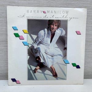 May include: Album cover for Barry Manilow's "I Wanna Do It With You." The cover features a photo of a man in a white suit. The album title and artist's name are printed in a script font. Colorful geometric shapes are scattered around the image.