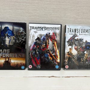 Op de afbeelding: Drie dvd-hoesjes van de Transformers-filmreeks. De hoesjes tonen afbeeldingen van robots en de filmtitels. De eerste hoes heet "Transformers", de tweede "Transformers: Dark of the Moon" en de derde "Transformers: Age of Extinction".