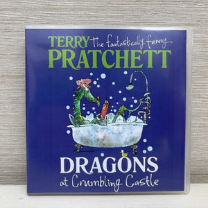 May include: A book cover with a blue background featuring the title "DRAGONS at Crumbling Castle" and the author's name, Terry Pratchett. An illustration shows a green dragon in a bathtub with bubbles. The book is in a clear protective sleeve.