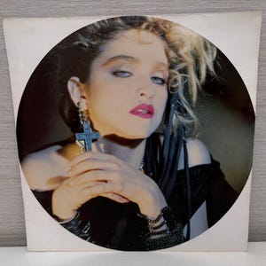 May include: Album cover featuring a woman with blonde hair and blue eyes, wearing a black top and cross earrings. The image is set against a dark background, with the woman's face in the center.
