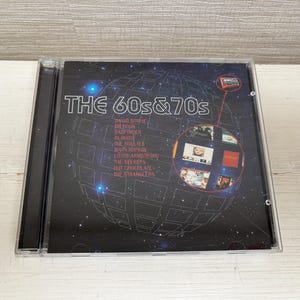 Puede incluir: Una caja de CD con el título "THE 60s & 70s" en blanco. La portada es negra con un diseño de globo geométrico y nombres de artistas como David Bowie y Blondie. El CD está en una funda de plástico transparente.