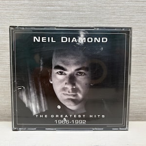 Puede incluir: Una caja de CD con una imagen en blanco y negro de Neil Diamond. El texto "NEIL DIAMOND" está sobre "THE GREATEST HITS" y los años "1966-1992". La caja está en una funda de plástico transparente.