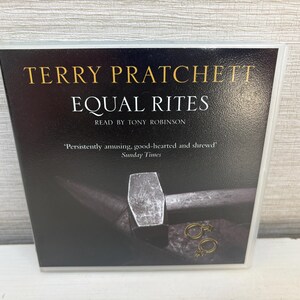 May include: A book cover for "Equal Rites" by Terry Pratchett, read by Tony Robinson. The cover features a dark background with the title in white and gold lettering. A hammer and other tools are visible.