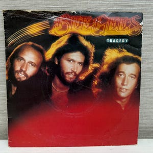 Puede incluir: Portada de álbum de vinilo vintage de "Tragedy" de los Bee Gees. La portada presenta a tres hombres con pelo largo, sobre un fondo rojo y negro. El nombre de la banda está en letras doradas, con el título del álbum debajo.