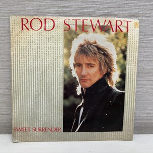 May include: A vintage album cover for Rod Stewart. The cover features a portrait of Stewart with blonde hair and a black jacket. The text "ROD STEWART" is in red at the top, and "SWEET SURRENDER" is at the bottom.