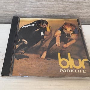 Op de afbeelding: Een muziek-cd-hoesje met de albumhoes van Blur's Parklife. De hoes toont twee hazewinden die op een baan rennen, één zwart en één bruin, beide met een muilkorf om. Het woord blur staat in grote gele letters op de hoes gedrukt met PARKLIFE eronder.