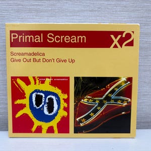 Puede incluir: Una caja de CD con el texto "Primal Scream" en rojo sobre un fondo amarillo. El título del álbum es "Screamadelica Give Out But Don't Give Up". La portada presenta un diseño solar y una bandera de neón.