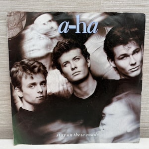 Pode incluir: Capa do álbum "Stay on These Roads" de a-ha. A capa apresenta uma imagem em tons sépia dos membros da banda com o nome da banda em azul claro no topo. O título do álbum está na parte inferior.