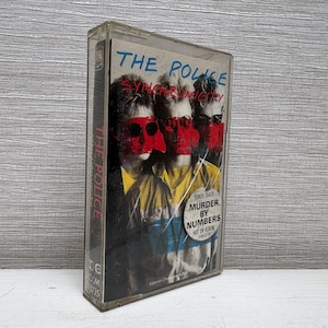 May include: A vintage cassette tape of The Police's album, Synchronicity, in a clear plastic case. The album cover features the band members with red masks. The bonus track "Murder by Numbers" is also noted on the cover.
