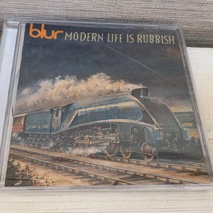 Op de afbeelding: Een doorzichtig plastic CD-doosje met de albumhoes van Blur, Modern Life Is Rubbish. De afbeelding toont een donkerblauwe stoomlocomotief die over treinsporen rijdt onder een bewolkte lucht, met de bandnaam en de albumtitel duidelijk zichtbaar.