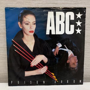 Può includere: Copertina dell'album "Poison Arrow" degli ABC con una donna che tiene delle frecce rosse. Il nome della band è in grandi lettere bianche con tre stelle. La copertina ha uno sfondo blu scuro.
