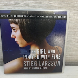 May include: A book cover for "The Girl Who Played with Fire" by Stieg Larsson. The cover features a woman's profile against a dark blue background. Text includes the title, author's name, and "Read by Martin Wenner."