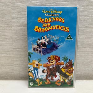 Op de afbeelding: Een VHS-cassette van de Disney-film "Bedknobs and Broomsticks". De hoes heeft een blauwe hemelachtergrond met de titel van de film in geel en oranje. De illustratie toont personages op een bed en geanimeerde wezens.