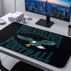 May include: Black desk mat featuring the words "PHILADELPHIA FOOTBALL" in teal, with a teal and white eagle graphic holding a football. The mat is rectangular and sits on a white desk, with a computer monitor and other office supplies visible.