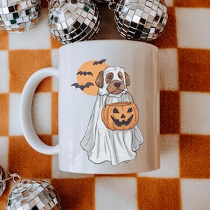 May include: White ceramic mug featuring a Halloween-themed illustration. The design includes a dog dressed as a ghost, holding a jack-o'-lantern, with bats and a full moon in the background. The mug has a curved handle and is set against a checkered orange and white background.