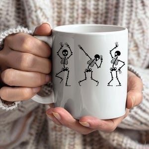 May include: White ceramic mug featuring three black skeleton illustrations in various dance poses. The skeletons have detailed rib cages, skulls, and limbs. The mug has a standard handle and a glossy finish, perfect for Halloween or spooky season.