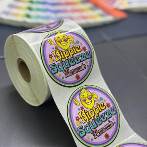 May include: A roll of circular adhesive labels featuring a yellow lemon character wearing sunglasses and making a peace sign. The design includes the text Hippie Squeeze Lemonade in blue and yellow, surrounded by small colorful flower icons on a purple background.