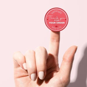 Pode incluir: Um autocolante redondo vermelho com texto branco que diz "Handmade, Thank you for your order, with love." O autocolante é segurado por um dedo contra um fundo rosa claro. O autocolante tem uma borda branca.