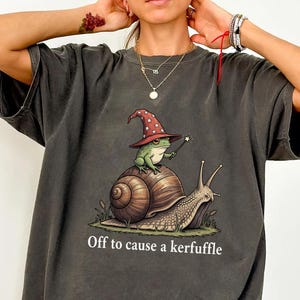 Könnte beinhalten: Dunkelgraues T-Shirt mit einer skurrilen Illustration eines Frosches mit Zauberhut auf einer Schnecke. Der Text "Off to cause a kerfuffle" ist darunter gedruckt. Das T-Shirt hat kurze Ärmel und eine lockere Passform.